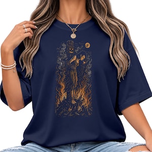 May include: A navy blue t-shirt featuring a graphic illustration of a person tied to a wooden post surrounded by orange flames. Above the person is a small jack-o'-lantern face on the post and a crescent moon in the dark, textured background.