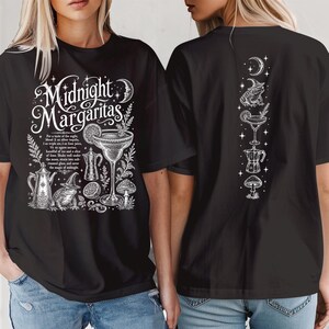 May include: A black t-shirt featuring white graphic illustrations and text. The front displays the text Midnight Margaritas with a cocktail glass, a toad wearing a witch hat, a coffee pot, and a mushroom. The back features a vertical design.