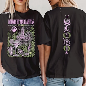 May include: A black t-shirt featuring a graphic print with the text "MIDNIGHT MARGARITAS EST. 1998". The design includes a house, a margarita glass, a frog, a coffee maker, and citrus slices. The back shows a vertical column of icons.