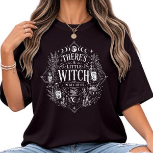 May include: A black short-sleeved t-shirt featuring a white graphic print of a cauldron, crystals, candles, and potion bottles arranged in a diamond shape. The text reads, "THERE'S A LITTLE WITCH IN ALL OF US," surrounded by moon phases and botanical elements.