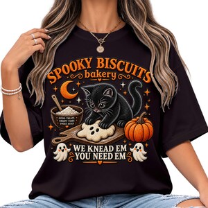 May include: A black t-shirt featuring a graphic of a black kitten baking on a wooden board with a pumpkin, a bowl of flour, and two small ghosts. The text reads SPOOKY BISCUITS bakery, GOOD TREATS, CREEPY VIBES, SWEET BITES, WE KNEAD EM YOU NEED EM.