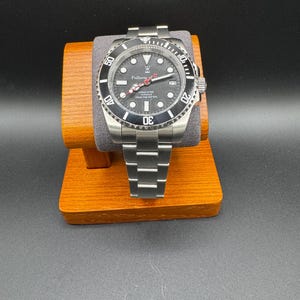 Puede incluir: Un reloj de buceo de acero inoxidable con esfera negra, bisel giratorio y brazalete de eslabones plateados. La esfera presenta marcadores de hora circulares blancos, una ventana de fecha en la posición de las tres y el texto Fullness of Time.