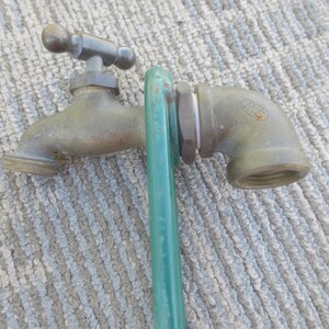May include: A brass-colored metal water spigot with a T-shaped handle and a threaded spout. A green metal rod is positioned vertically against the side of the spigot body. The spigot features a curved elbow fitting with the letters MS stamped on it.
