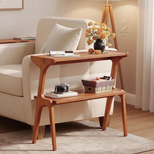 May include: A two-tiered wooden console table with a warm brown finish. The top surface holds a black vase with orange flowers and a small book. The lower shelf features a vintage-style camera resting on a book next to a rectangular woven basket.