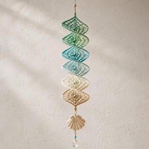 Puede incluir: Un móvil en espiral colgante que presenta seis adornos de ganchillo en forma de diamante en un degradado de tonos verdes, azules y beige. La estructura está suspendida por un hilo y rematada en la parte inferior con una concha de ganchillo.
