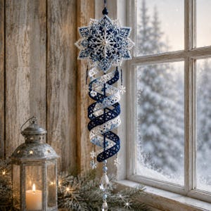 Könnte beinhalten: Ein hängendes Ornament mit einer großen, filigranen, weiß-dunkelblauen Häkelschneeflocke an der Spitze, verbunden mit einer spiralförmigen, bandartigen Häkelstruktur in passenden Farben, akzentuiert durch kleine Schneeflockenanhänger und klare Kristalltropfen.