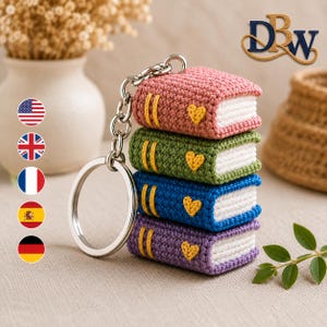 May include: A crochet keychain featuring a stack of four miniature books in pink, green, blue, and purple. Each book has white pages, two yellow vertical lines on the spine, and a small yellow heart on the cover, attached to a silver ring.
