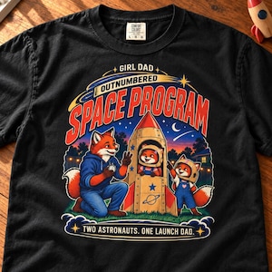 Peut inclure: Un t-shirt noir avec un motif de trois renards sur le thème de l'espace. Le texte indique GIRL DAD OUTNUMBERED SPACE PROGRAM et TWO ASTRONAUTS. ONE LAUNCH DAD. Le design inclut une fusée, des étoiles et un croissant de lune nocturne.