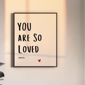 Può includere: Una stampa artistica da parete rettangolare in una cornice scura con il testo nero "YOU ARE SO LOVED" in un carattere sans-serif. Sotto il testo, la citazione "JOHN 3:16" è stampata in un carattere più piccolo, con un piccolo cuore rosso.