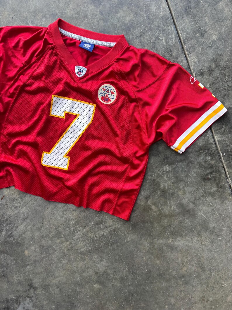 May include: A cropped red football jersey with the number 7 in white and a yellow outline. The jersey has a V-neck and white and yellow stripes on the sleeves. A team logo is visible on the chest.