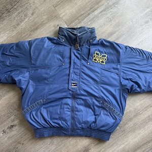 May include: A navy blue Nike puffer jacket featuring a high collar, a full-length front zip, and long sleeves. The chest displays a yellow embroidered Michigan block M logo and a small Nike swoosh logo on a rectangular patch above the zip.