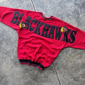 May include: Red sweatshirt with black lettering that reads "Blackhawks" with two embroidered blackhawk logos on the front.