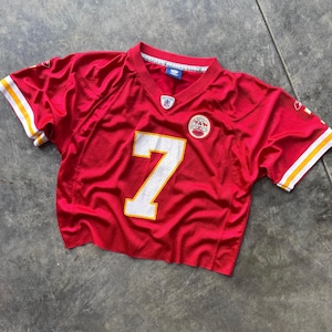 May include: A cropped red football jersey with the number 7 in white. The jersey has white and yellow stripes on the sleeves and a team logo patch on the chest. The jersey is made of a shiny, mesh-like material.