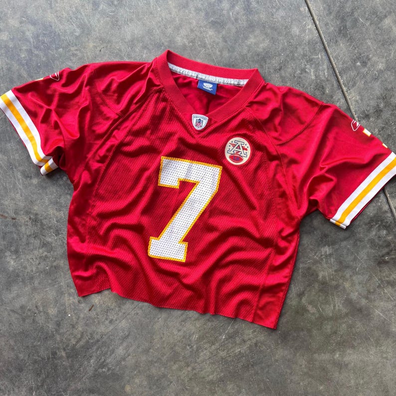 May include: A cropped red football jersey with the number 7 in white. The jersey has white and yellow stripes on the sleeves and a team logo patch on the chest. The jersey is made of a shiny, mesh-like material.