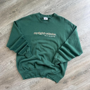 May include: A dark green crewneck sweatshirt featuring the text wright state RAIDERS embroidered in white and gold on the chest. The garment has long sleeves with ribbed cuffs and a ribbed neckline, with a Jerzees brand tag visible at the collar.
