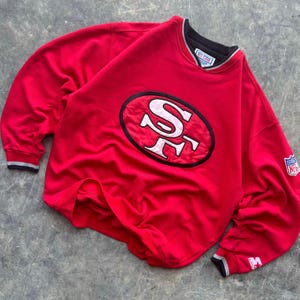 May include: Red sweatshirt with a large embroidered San Francisco 49ers logo on the front. The sweatshirt has a black and white striped trim around the neck and cuffs. The NFL logo is on the right sleeve.