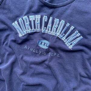 May include: Navy blue tank top with embroidered "NORTH CAROLINA" in light blue arched across the chest. Below the text is an oval with "XXL" and the words "ATHLETIC DEPT." are embroidered below.
