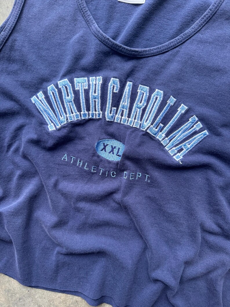 May include: Navy blue tank top with embroidered "NORTH CAROLINA" in light blue arched across the chest. Below the text is an oval with "XXL" and the words "ATHLETIC DEPT." are embroidered below.