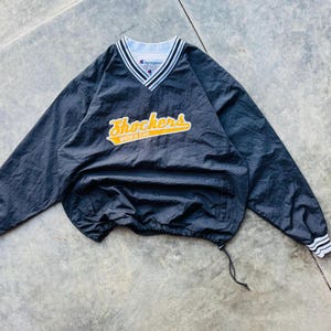 May include: Black Champion pullover windbreaker with a V-neck collar and the word "Shockers" in yellow and white lettering. The windbreaker has white and black striped cuffs and collar trim.