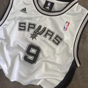 Puede incluir: Una camiseta de baloncesto de malla blanca con el logotipo de los San Antonio Spurs y el n&uacute;mero 9 en letras negras y plateadas. La prenda sin mangas incluye ribetes negros y plateados en el cuello y las sisas, con un logotipo de Adidas y NBA.
