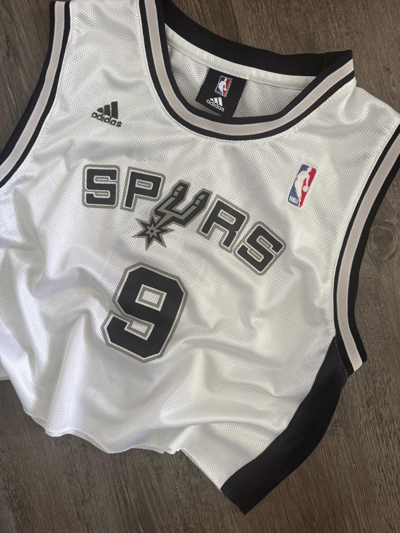 Puede incluir: Una camiseta de baloncesto de malla blanca con el logotipo de los San Antonio Spurs y el n&uacute;mero 9 en letras negras y plateadas. La prenda sin mangas incluye ribetes negros y plateados en el cuello y las sisas, con un logotipo de Adidas y NBA.