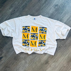 Peut inclure: Un t-shirt à manches courtes gris chiné clair avec un motif en grille composé de carrés jaunes portant la lettre M et des illustrations de carcajous bleu foncé. Le texte UNIVERSITY OF MICHIGAN est imprimé en jaune et bleu sous la grille.