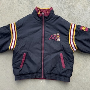 May include: A black zip-up jacket with maroon and gold accents. The jacket features a maroon collar and pocket trim, with gold and white stripes down the sleeves. A logo patch is on the chest and sleeve, with the letter "M" and a mascot.