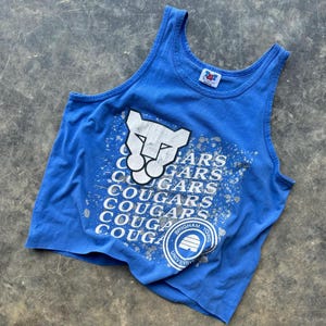 May include: A blue vest top with a white cougar head graphic and the word "COUGARS" repeated. The vest top also features the text "Brigham Young University" in a circular emblem.