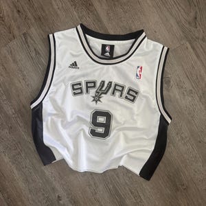 Puede incluir: Una camiseta de baloncesto blanca sin mangas con ribetes negros en el cuello y las sisas. El frente presenta el texto Spurs con un logotipo de espuela, el n&uacute;mero 9, un logotipo de Adidas y un logotipo de la NBA. Los lados son de malla negra.