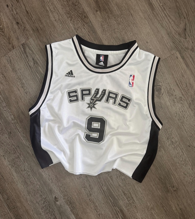 Puede incluir: Una camiseta de baloncesto blanca sin mangas con ribetes negros en el cuello y las sisas. El frente presenta el texto Spurs con un logotipo de espuela, el n&uacute;mero 9, un logotipo de Adidas y un logotipo de la NBA. Los lados son de malla negra.