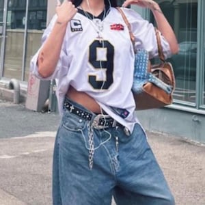 May include: White jersey with the number 9 in black, paired with light wash denim jeans. A black belt with silver accents and a chain is visible. The outfit is accessorized with a brown purse and sunglasses.
