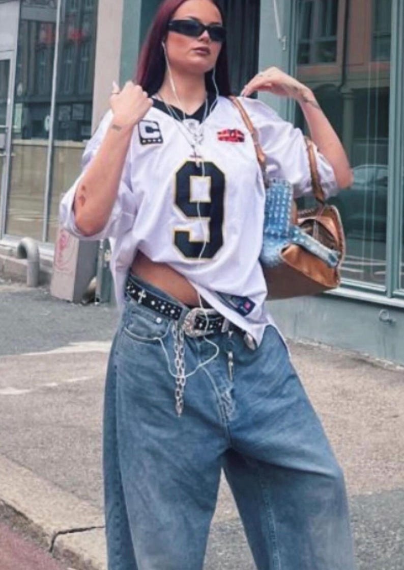 May include: White jersey with the number 9 in black, paired with light wash denim jeans. A black belt with silver accents and a chain is visible. The outfit is accessorized with a brown purse and sunglasses.