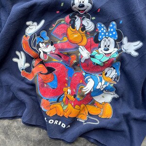 Puede incluir: Una camiseta azul marino con un gr&aacute;fico colorido de Mickey Mouse, Minnie Mouse, Goofy, Pluto y Donald Duck. El gr&aacute;fico incluye la palabra "Florida" y el s&iacute;mbolo de copyright de Disney.