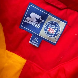 May include: A red and yellow Starter brand jacket with a blue label that says "Starter" and "Officially Licensed NFL Product". The label also has a star and an NFL logo. The jacket has a size tag that says "XL" and "Made in Philippines".