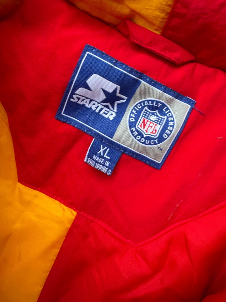 May include: A red and yellow Starter brand jacket with a blue label that says "Starter" and "Officially Licensed NFL Product". The label also has a star and an NFL logo. The jacket has a size tag that says "XL" and "Made in Philippines".