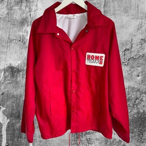May include: A red nylon jacket with a white zip and a white drawstring. The jacket has a patch on the left chest that says "HOME TEAM".