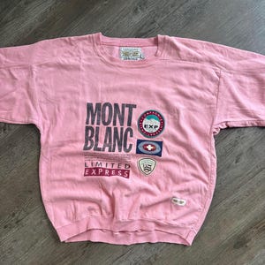 May include: A light pink crewneck sweatshirt featuring the text MONT BLANC LIMITED EXPRESS in dark gray. The front includes three circular and rectangular patches with mountain and Swiss-inspired graphics, plus a small rectangular brand label.