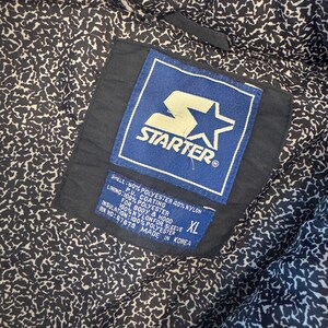 May include: Close-up of the inside of a black Starter jacket. The lining has a white and black pattern. A blue and white Starter label is visible, along with a size XL tag and fabric composition details.