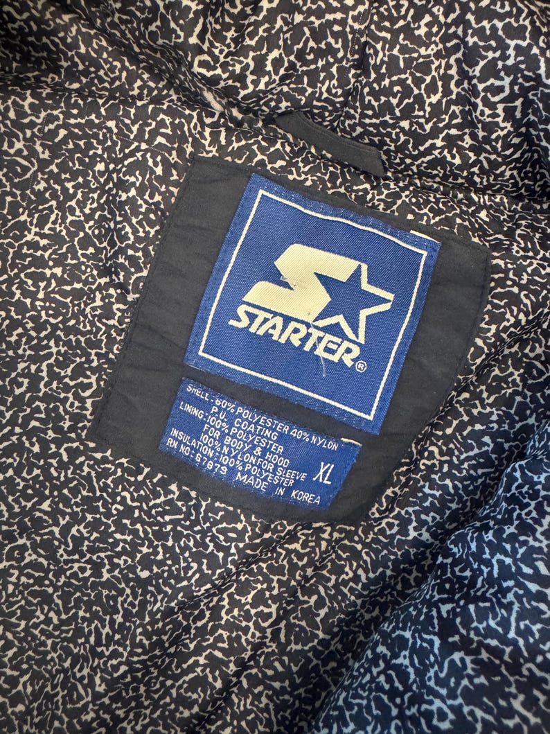 May include: Close-up of the inside of a black Starter jacket. The lining has a white and black pattern. A blue and white Starter label is visible, along with a size XL tag and fabric composition details.
