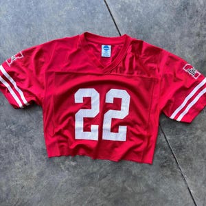 May include: A cropped red football jersey with the number 22 in white on the front. The jersey has white stripes on the sleeves and a V-neck. The NCAA logo is visible on the neckline.