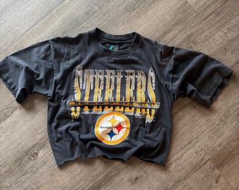 Puede incluir: Una camiseta recortada de color gris carbón con la palabra "STEELERS" en letras doradas y blancas. Debajo de la palabra "STEELERS" está la palabra "PITTSBURGH" en dorado. La camiseta también muestra el logotipo de los Steelers.