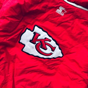 May include: Red Starter brand jacket with a white and black Kansas City Chiefs logo on the back. The logo is a stylized arrowhead with the letters KC inside.