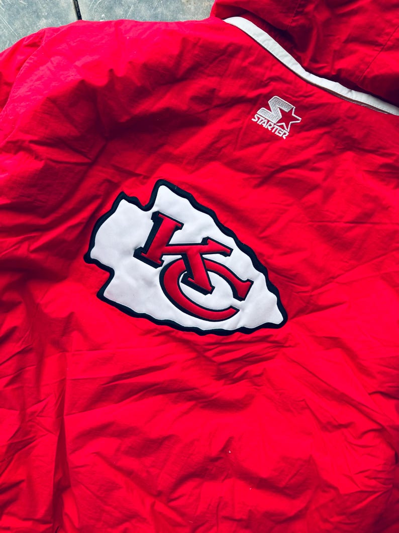 May include: Red Starter brand jacket with a white and black Kansas City Chiefs logo on the back. The logo is a stylized arrowhead with the letters KC inside.