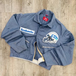 May include: A dusty blue jacket with a zip closure, featuring a Seahawks patch and the team name on the front. The jacket has two front pockets and a collar. The Seahawks logo is a white and blue helmet.