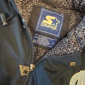 May include: Black Starter jacket with a zip closure and a light blue and white patterned interior lining. The jacket features the Starter logo on the zipper pull and a patch with the Starter logo and text. The jacket has an XL size tag.
