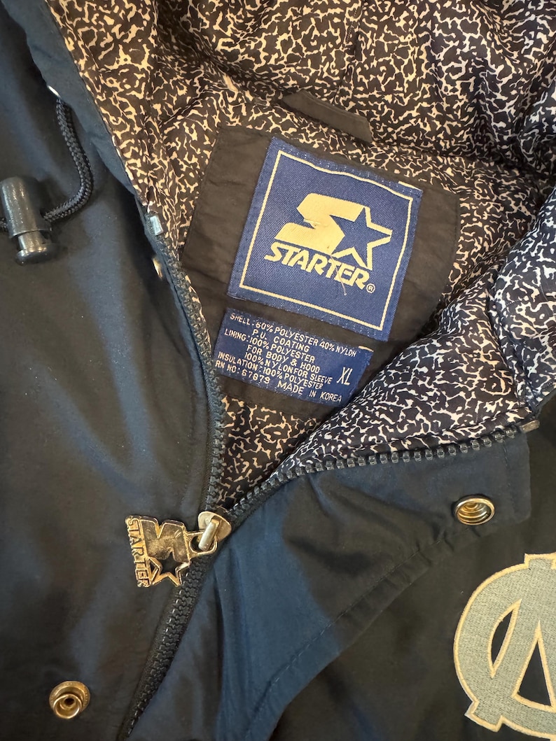 May include: Black Starter jacket with a zip closure and a light blue and white patterned interior lining. The jacket features the Starter logo on the zipper pull and a patch with the Starter logo and text. The jacket has an XL size tag.