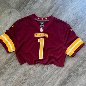May include: A burgundy short-sleeved jersey featuring the word COMMANDERS in yellow text above the number 1 in yellow with a white outline. The sleeves have yellow and white stripes, and the neckline displays three yellow stars and an NFL logo.