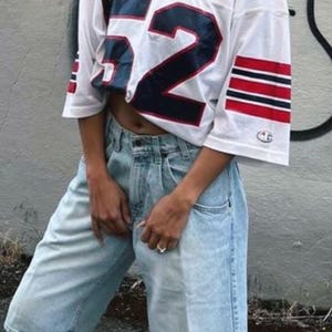 May include: White football jersey with the number 52 in navy blue and red stripes on the sleeves. The jersey is paired with light wash denim shorts. The outfit is casual and sporty.