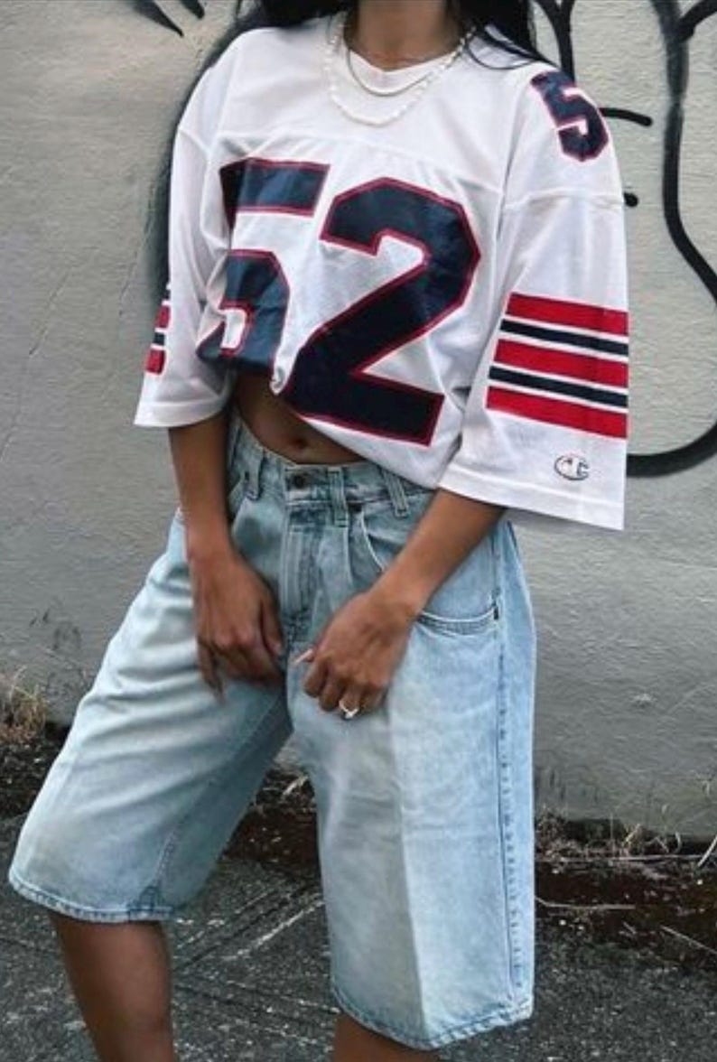 May include: White football jersey with the number 52 in navy blue and red stripes on the sleeves. The jersey is paired with light wash denim shorts. The outfit is casual and sporty.