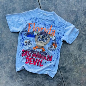May include: A light blue t-shirt with a white splatter paint design. The shirt features a cartoon Tasmanian Devil wearing a University of Florida football uniform. The text "University of Florida" and "Tasmanian Devil" are printed on the shirt.